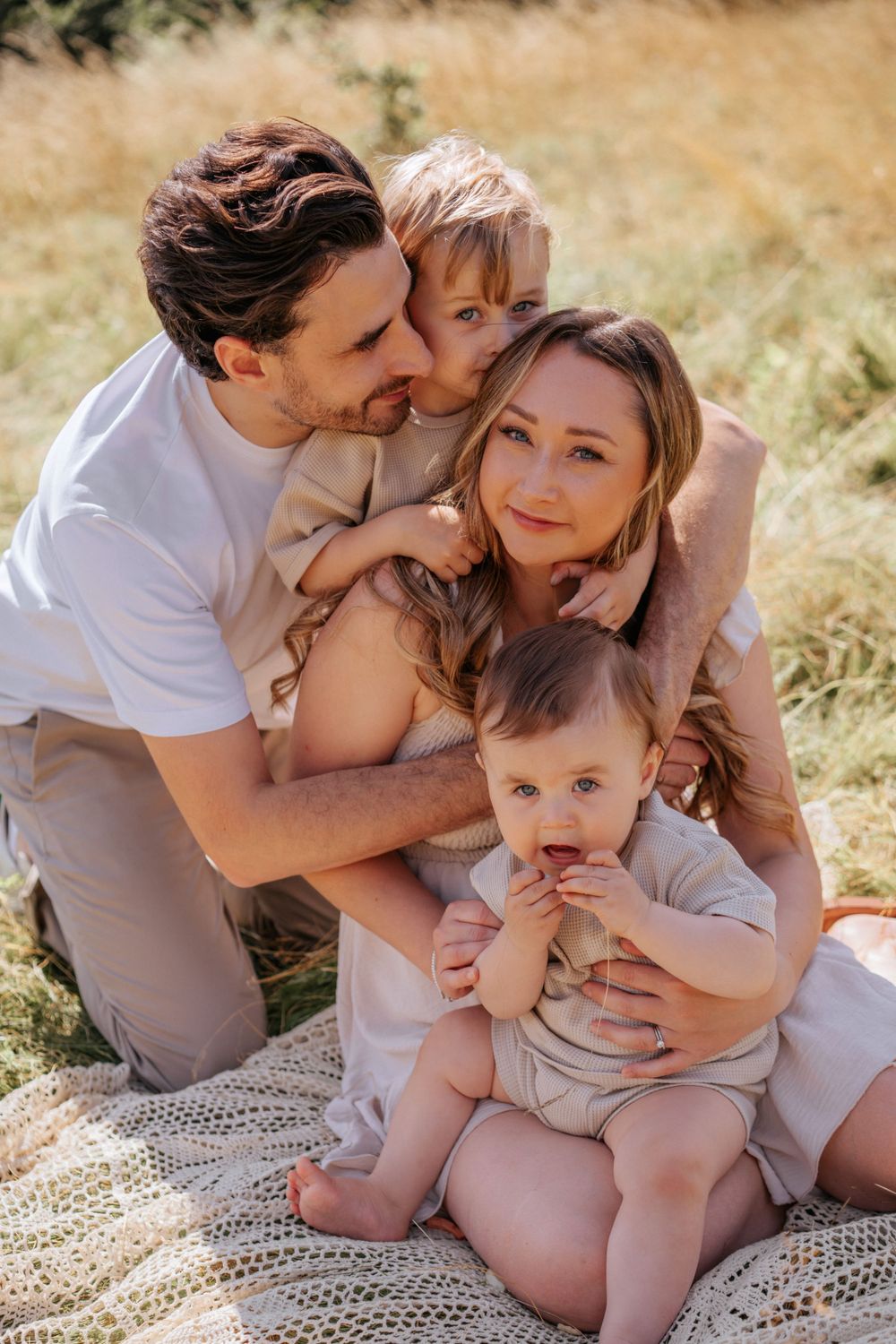 Family sits together on knitted blanket outdoors enjoying tender moment in golden summer field. Family Photoshoot.