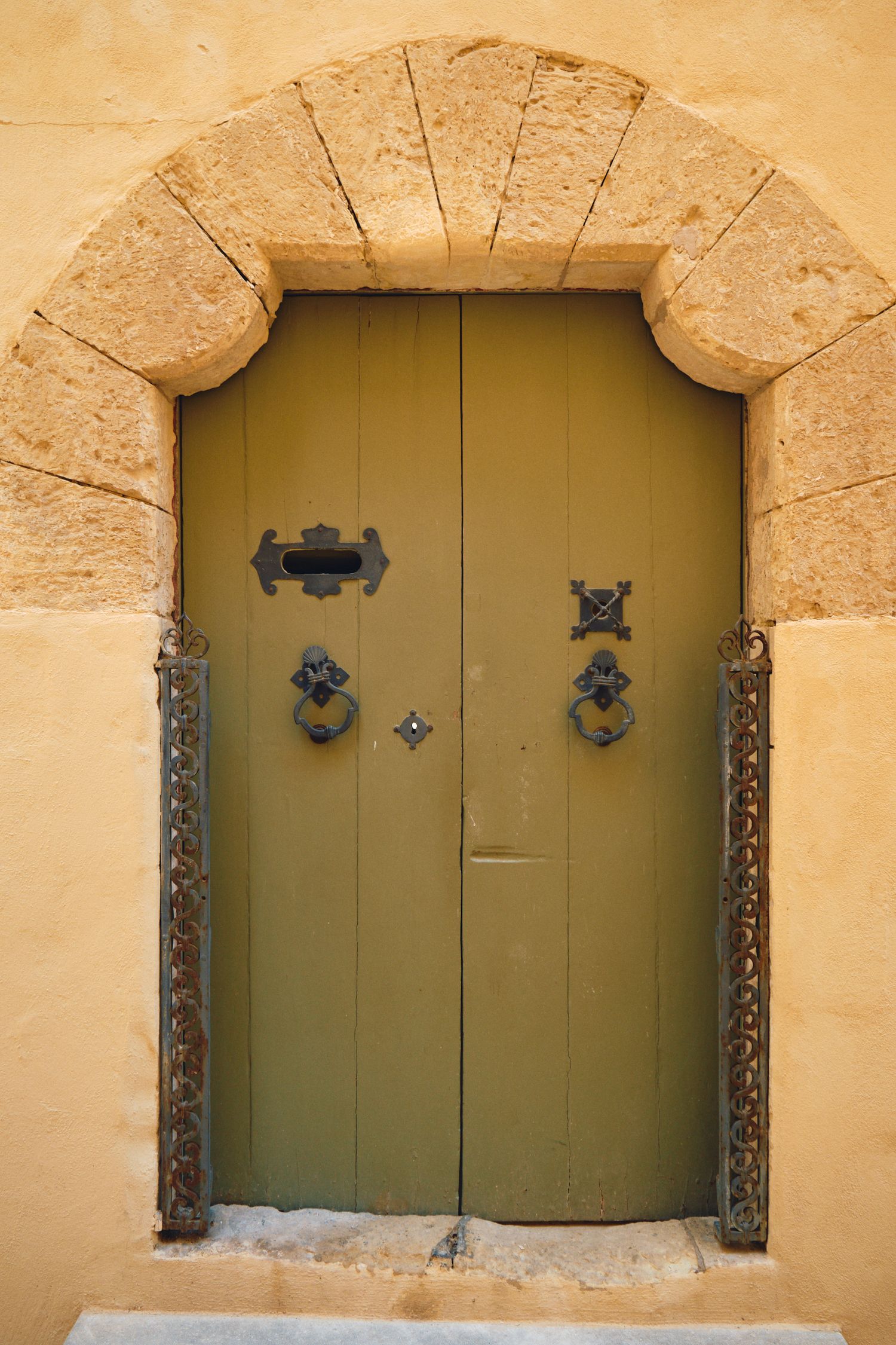 Rustic olive green double door with ornate metal hardware framed by a stone archway in Mediterranean architecture.