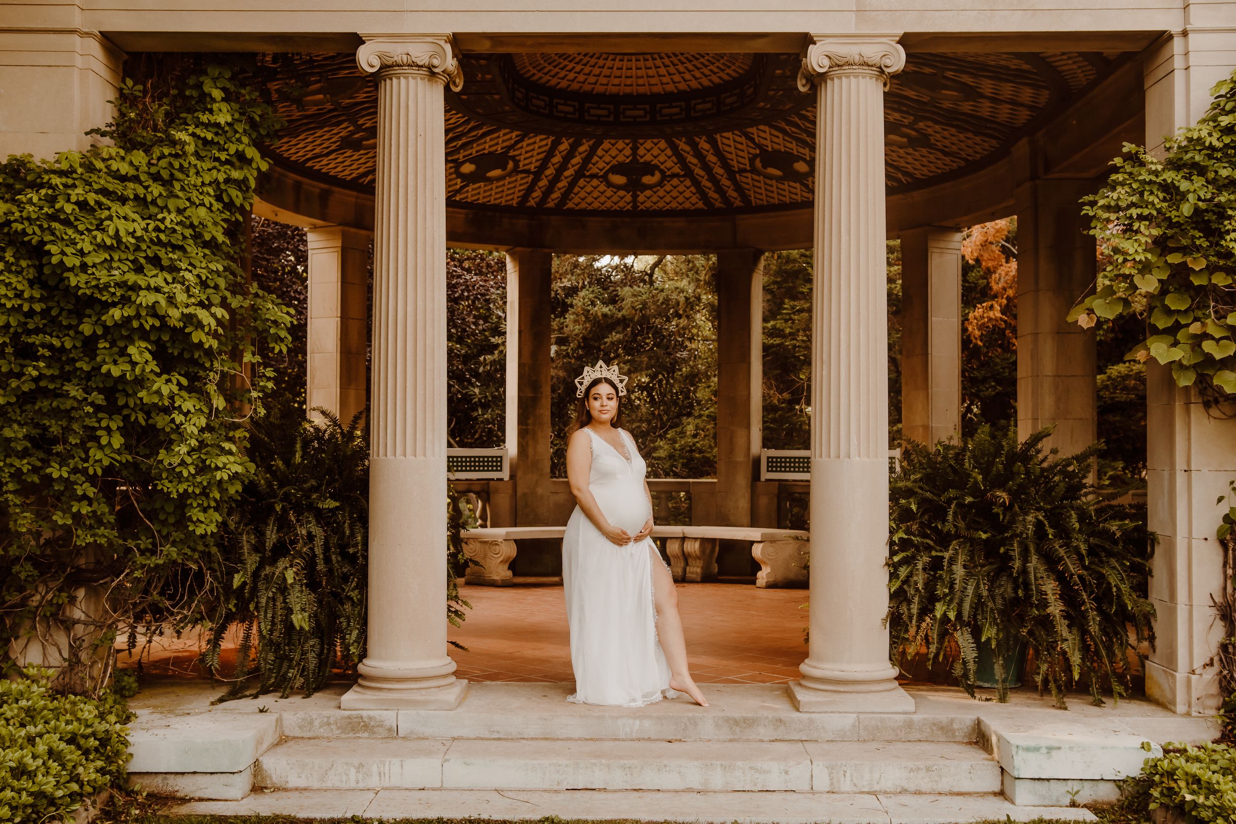 A figure in a white flowing dress stands gracefully in an elegant gazebo surrounded by lush greenery at golden hour in Waterford, CT.