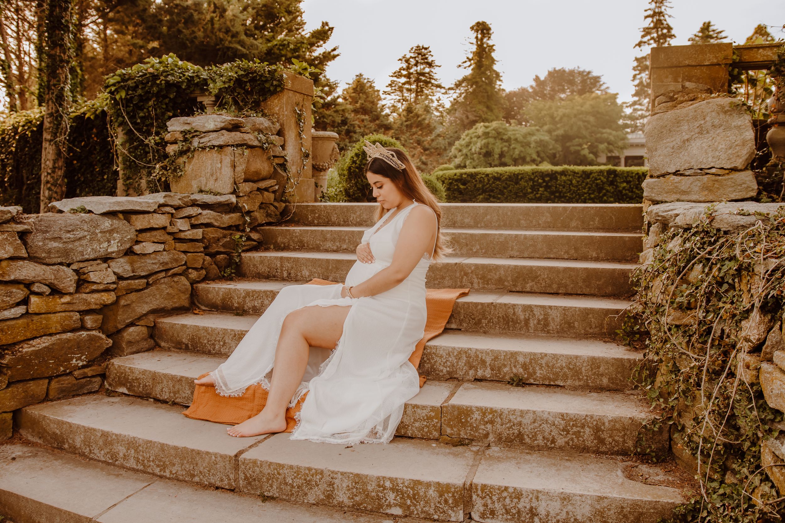 A mother in a flowing white dress sits gracefully on rustic stone steps at sunset in a garden setting in Waterford, Connecticut.