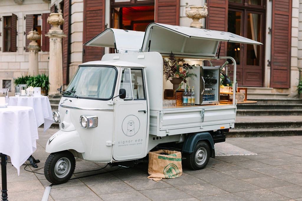 A vintage white three-wheeled coffee truck with open serving hatch parked in front of a historic building entrance.