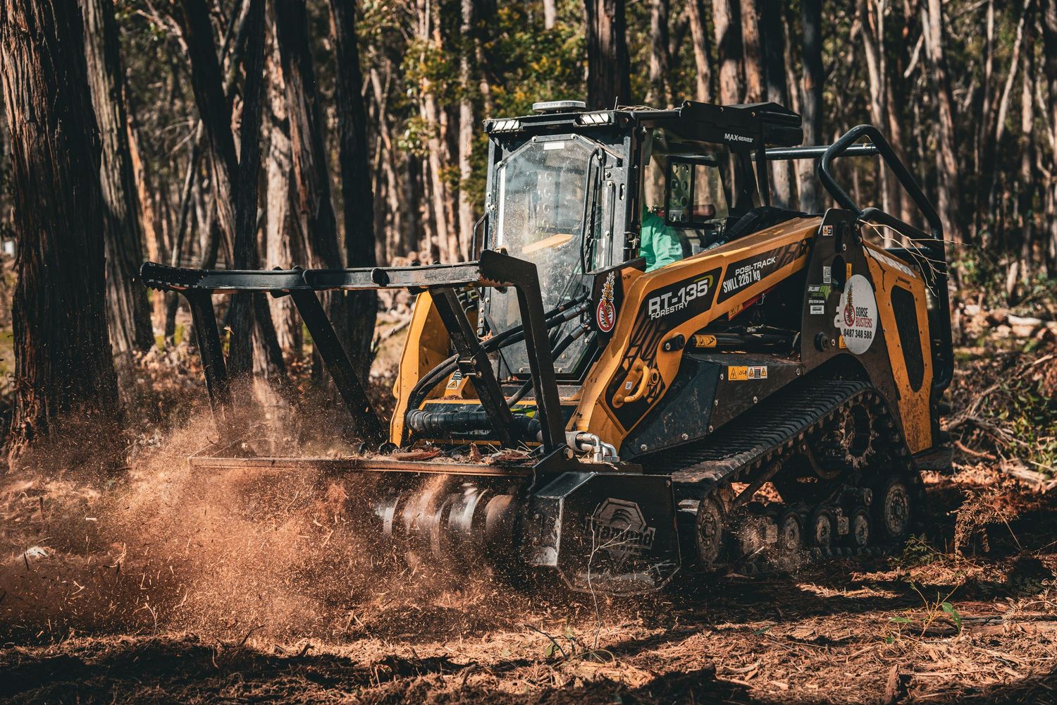 Orange compact track loader kicking up dirt while operating between trees in a wooded area.