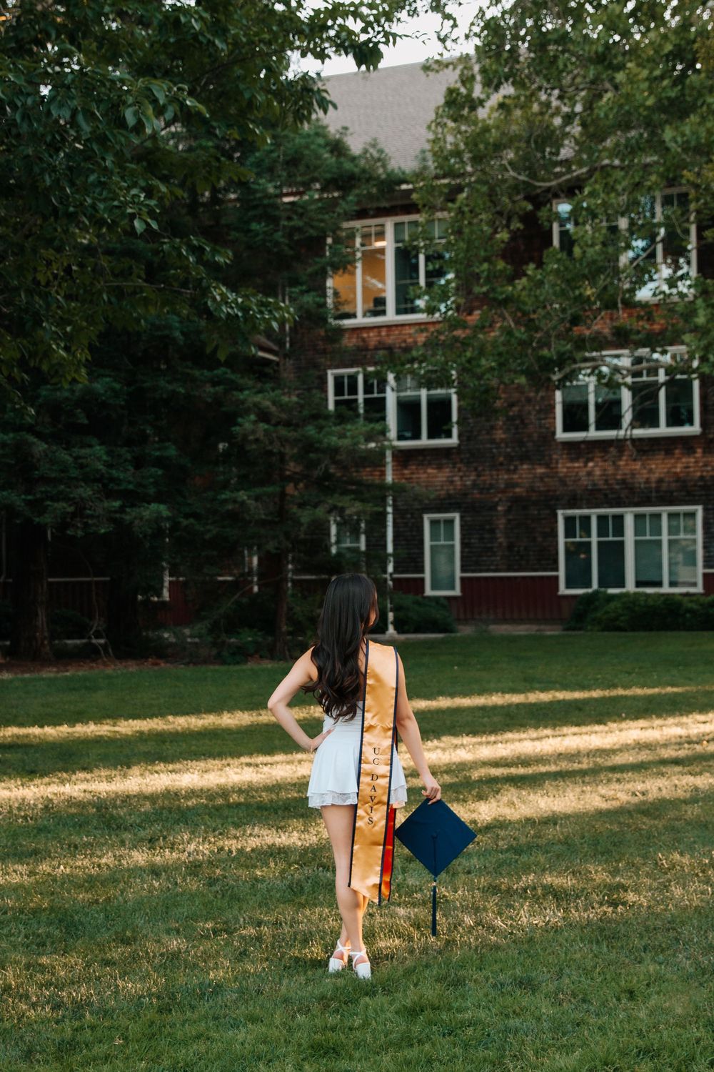 Graduate stands in golden sunlight on campus lawn wearing white dress and yellow sash while holding blue diploma.