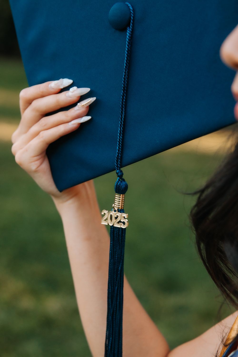 Close-up view of a navy blue graduation cap with a 2023 tassel charm and white manicured nails holding the corner.