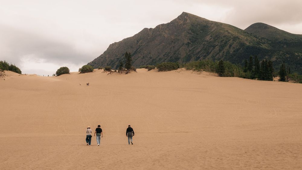 Three hikers traverse vast sand dunes beneath towering mountain peaks on a cloudy day.