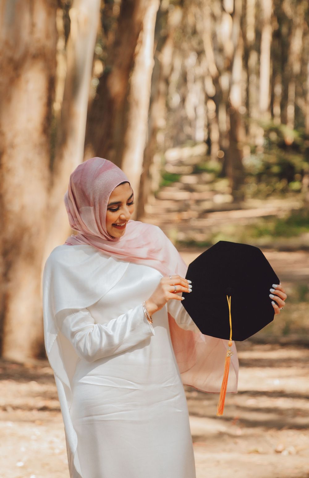 Person in white dress and pink hijab holding graduation cap along wooded path in warm sunlit forest setting.