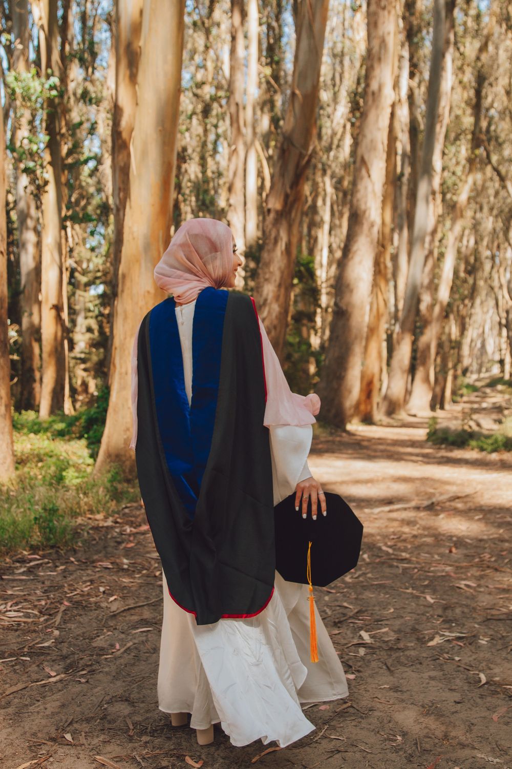 Person wearing a navy blue and black graduation gown walks along a eucalyptus tree-lined path in a forest setting.