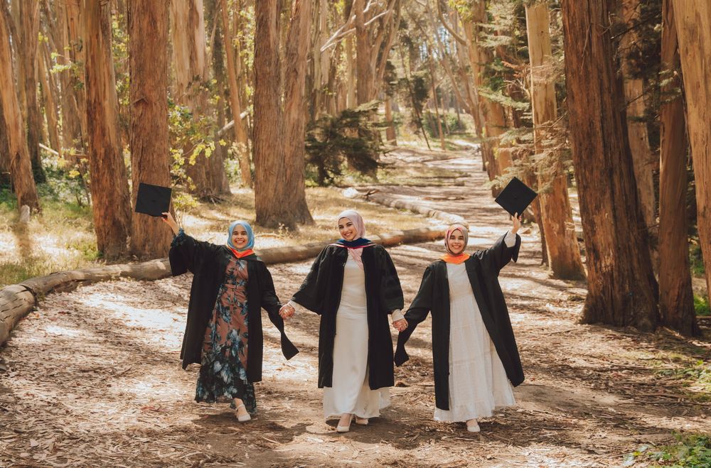Three people wearing graduation gowns hold hands while walking down a tree-lined path in a redwood forest.