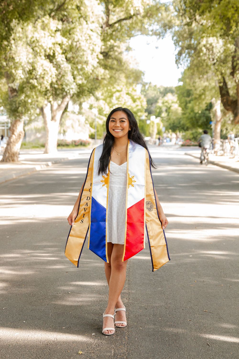 Graduate wearing a white dress and Filipino flag-inspired graduation stole walks down a tree-lined street.