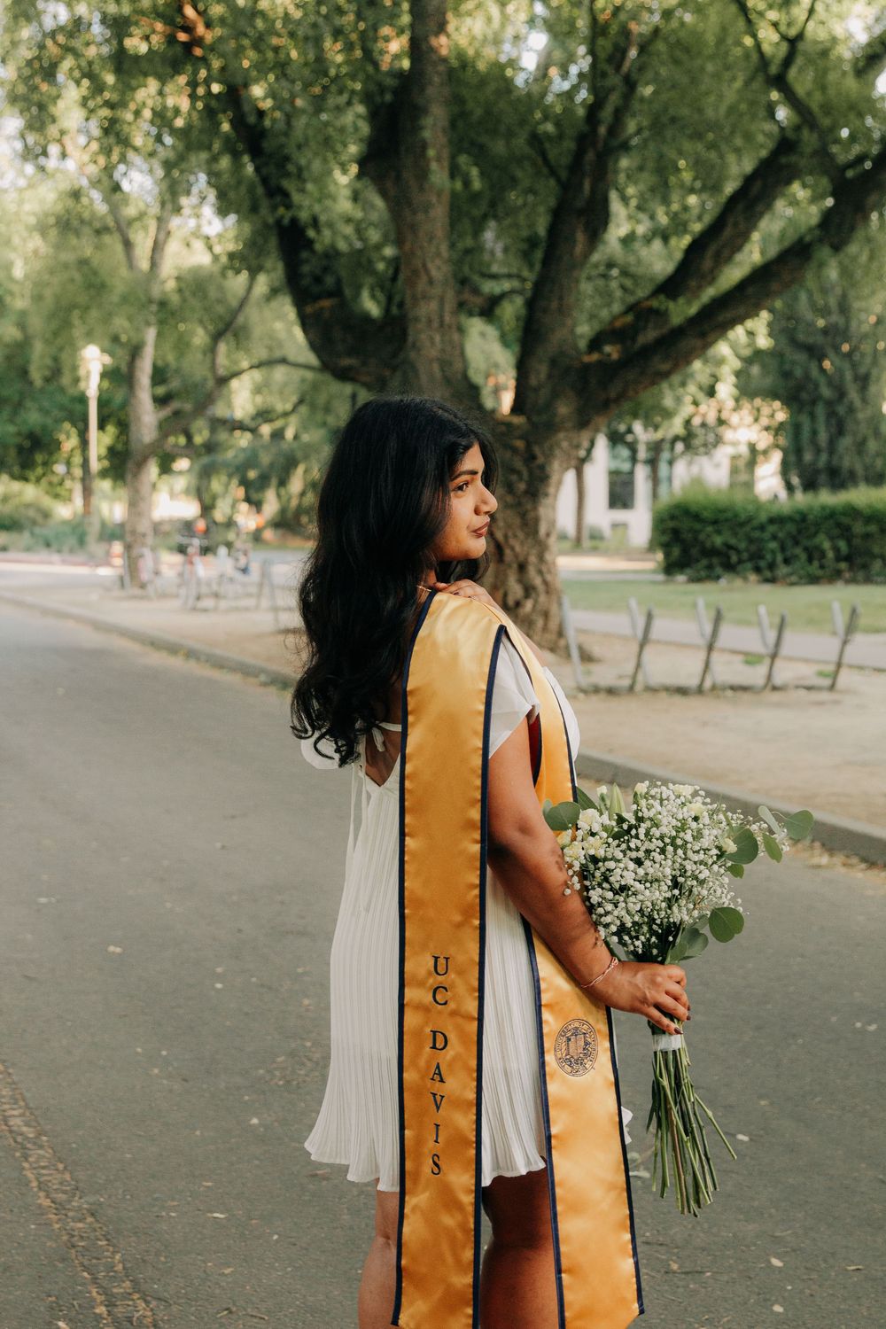 Graduate in white dress and gold sash holding a bouquet of flowers stands on a tree-lined path during graduation photoshoot.