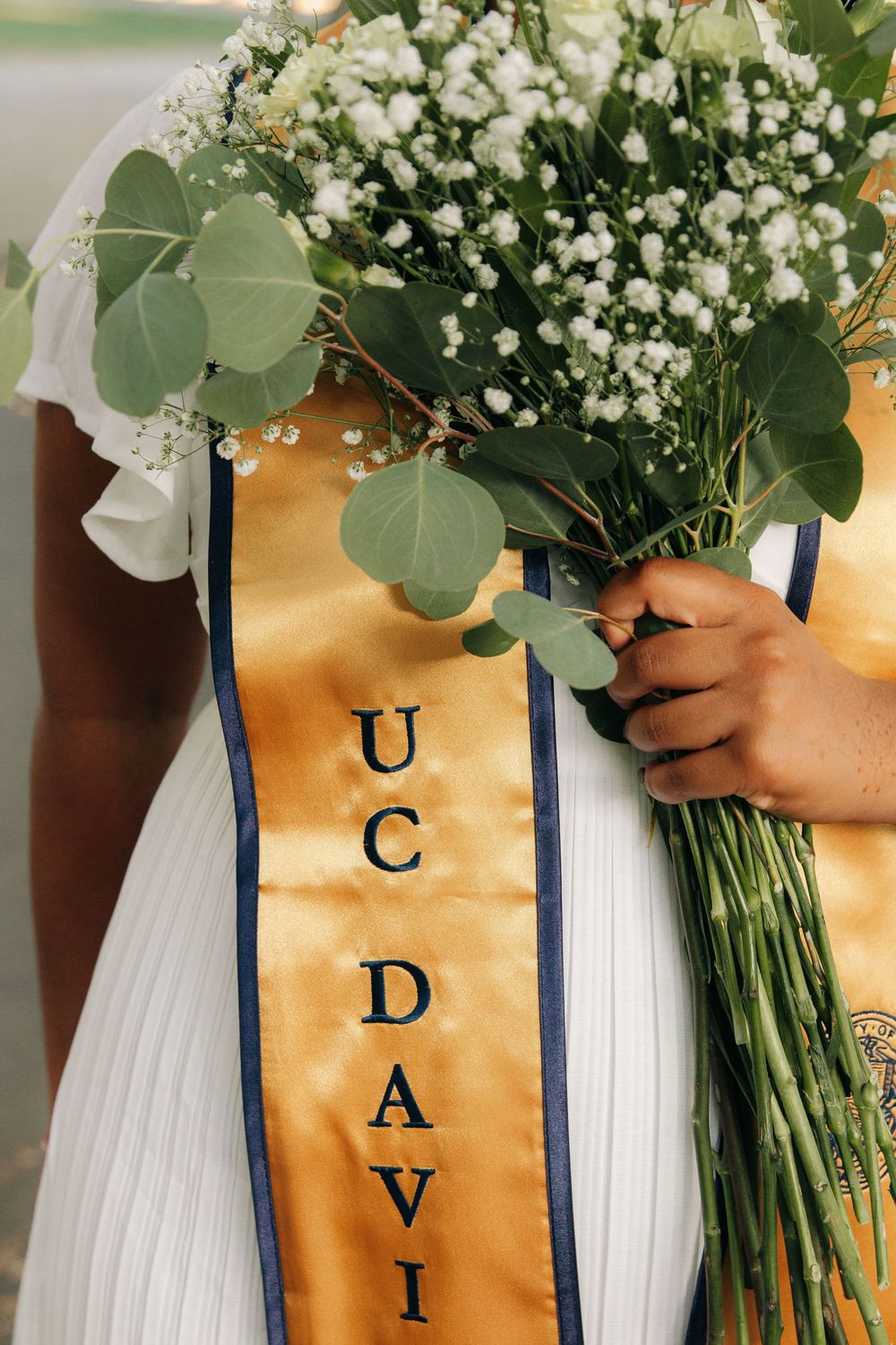 A bridal bouquet of baby's breath and eucalyptus leaves held against a white dress with a UC Davis sash.