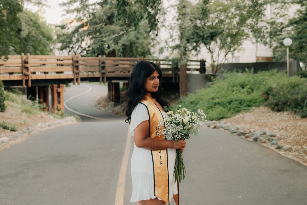 A person in a white dress holds a bouquet of wildflowers on a winding path with a wooden bridge in the background.