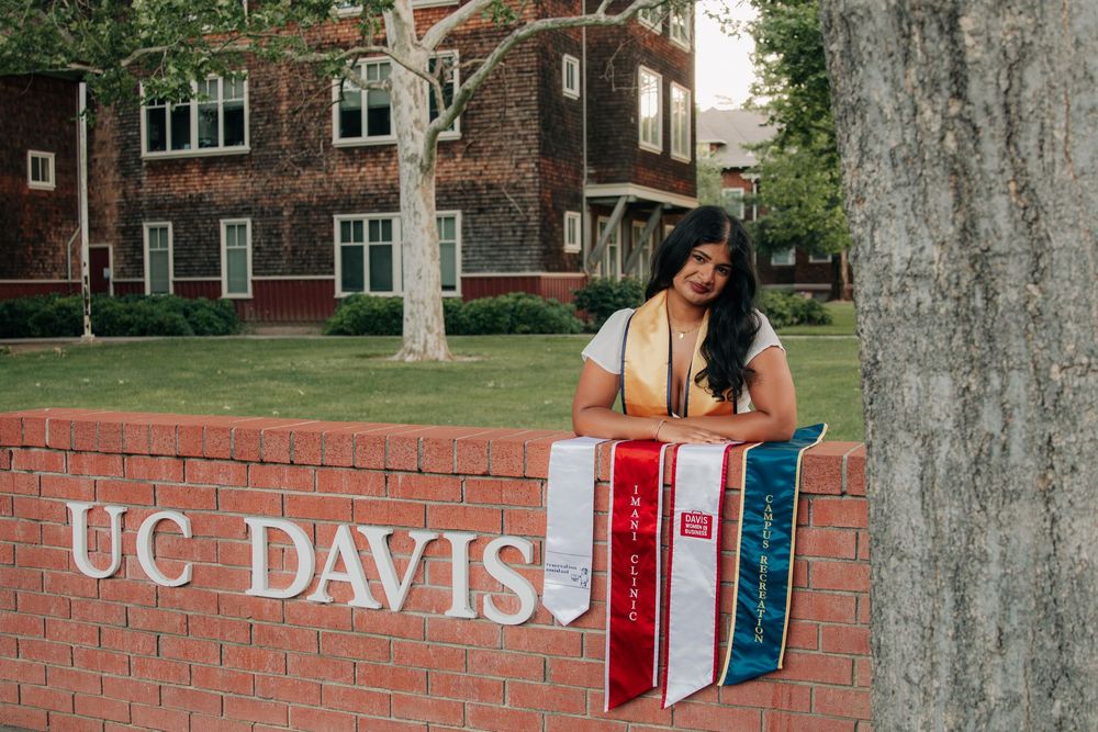 A graduate poses next to the UC Davis brick sign on campus with historic buildings visible in the background.