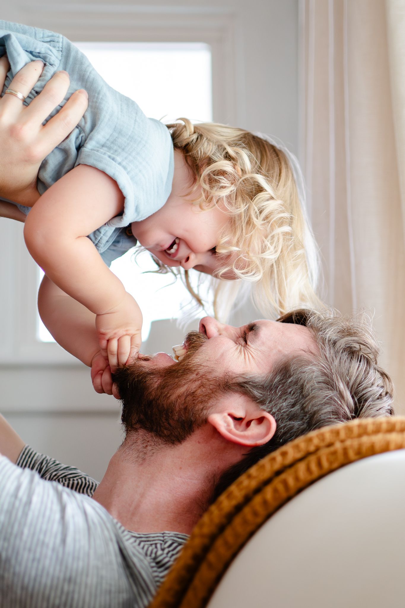 A dad holds his little girl up in the air, creating laughs for both of them during an in-home family photo shoot.
