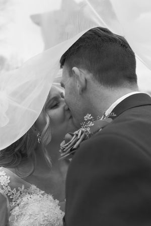 Black and white photo of bride and groom sharing a romantic kiss under a flowing wedding veil.