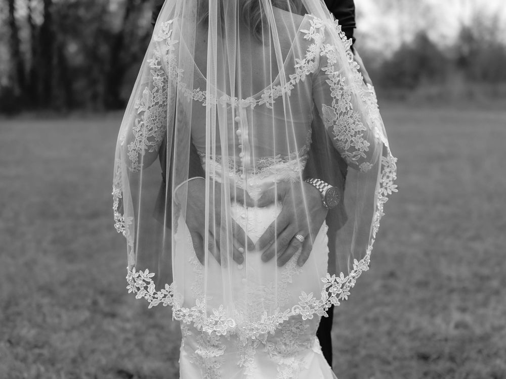 Black and white photo of a bride and groom embracing under a lace-trimmed wedding veil outdoors.