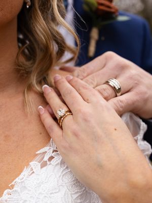 Close-up of bride and groom's hands showing gold wedding rings and diamond engagement ring against white lace dress.