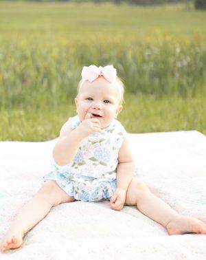 Baby in floral outfit lying on white blanket in grassy field during golden hour.