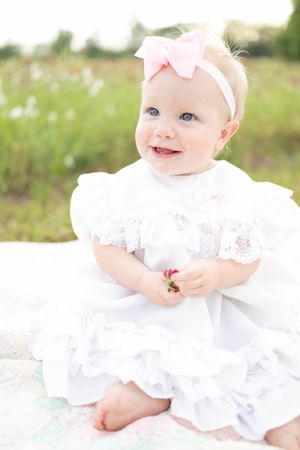 Baby wearing white lace dress sitting on blanket outdoors in sunlit field.