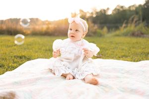 Baby in white dress lying on blanket outdoors with bubbles floating in golden sunlight.