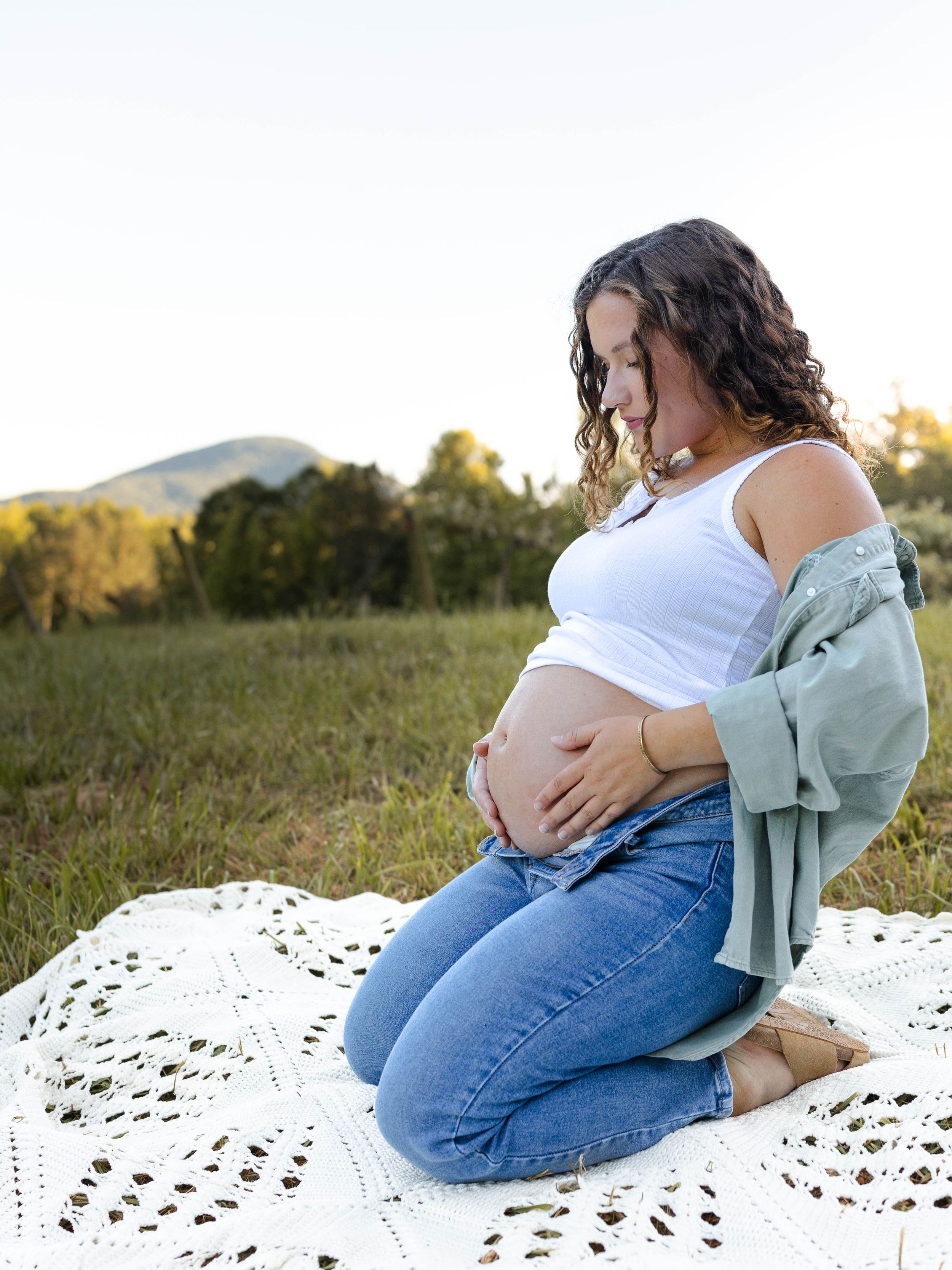 Maternity photos on white lace blanket in outdoor setting during sunset.