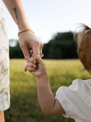 Adult holding a young child's hand in a sunny green field, wearing a gold bracelet and ring.