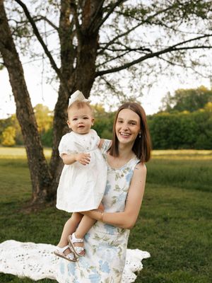 Young woman in floral dress holds smiling baby girl in white dress outdoors under trees at golden hour.