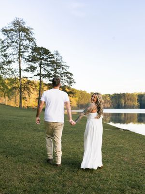 Couple holding hands walking toward a serene lake at golden hour, woman in white maxi dress with tattoos, man in white tee.
