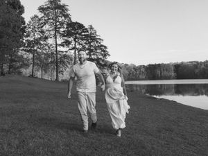 Black and white photo of a happy couple running and laughing together on a grassy lawn beside a serene lake.
