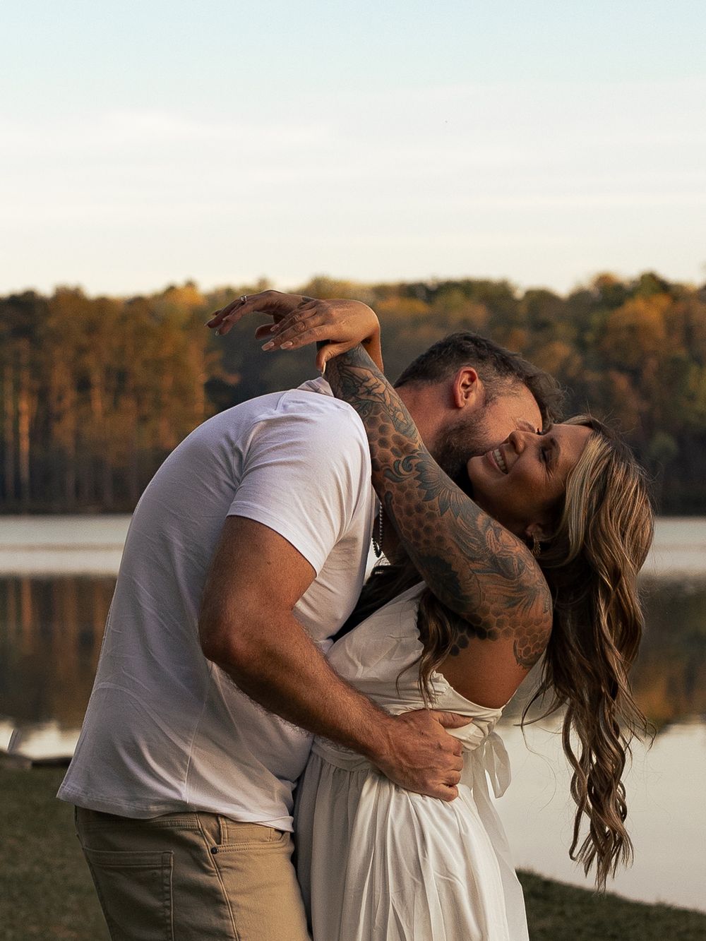 Couple embracing and kissing by a lake at golden hour, man with tattooed arms in white tee, woman in white dress.