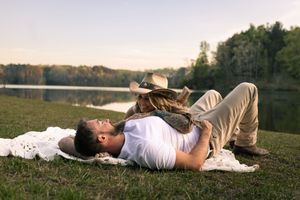 Romantic couple relaxing on a blanket by a lakeside at sunset, man lying down while woman leans over him.