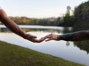 Two hands reaching toward each other near a serene lake at dusk, one arm adorned with tattoos.