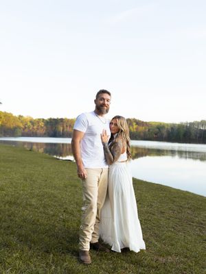 Couple posing together by a serene lake at sunset, woman in white dress, man in white shirt and khaki pants.
