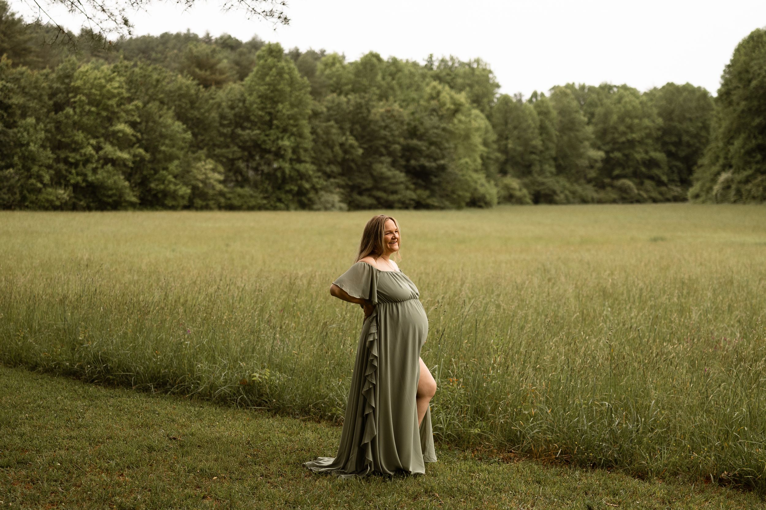 Pregnant woman in sage green maternity gown standing in lush green field with forest backdrop.
