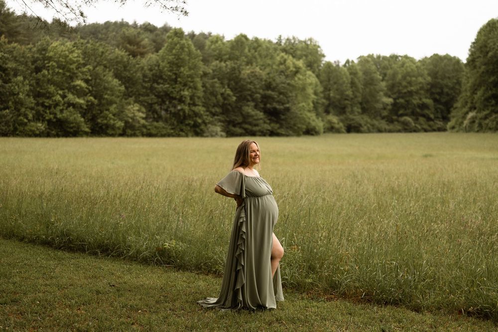 Pregnant woman in sage green maternity gown standing in lush green field with forest backdrop.