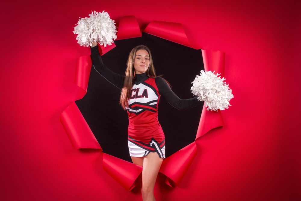 Cheerleader in red and white uniform poses with pom poms through torn red paper background for dramatic effect.