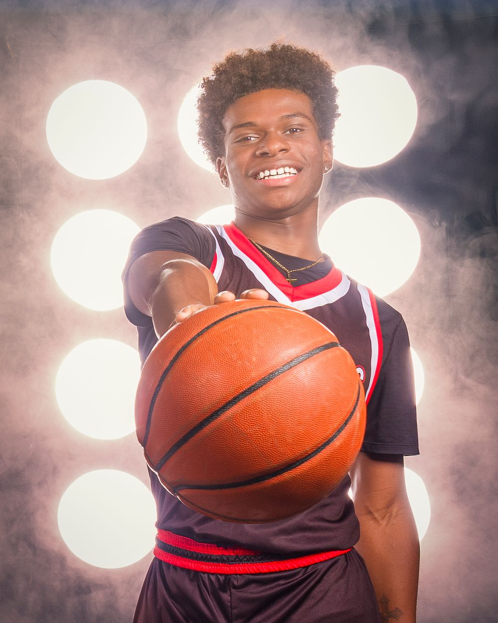High school basketball player poses with ball against white spotlights in dramatic sports portrait.