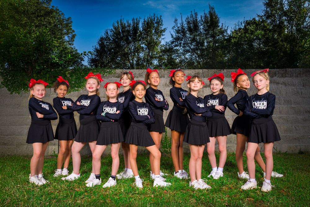 A cheerleading squad in black uniforms and red bows poses together outdoors against a gray wall and trees.