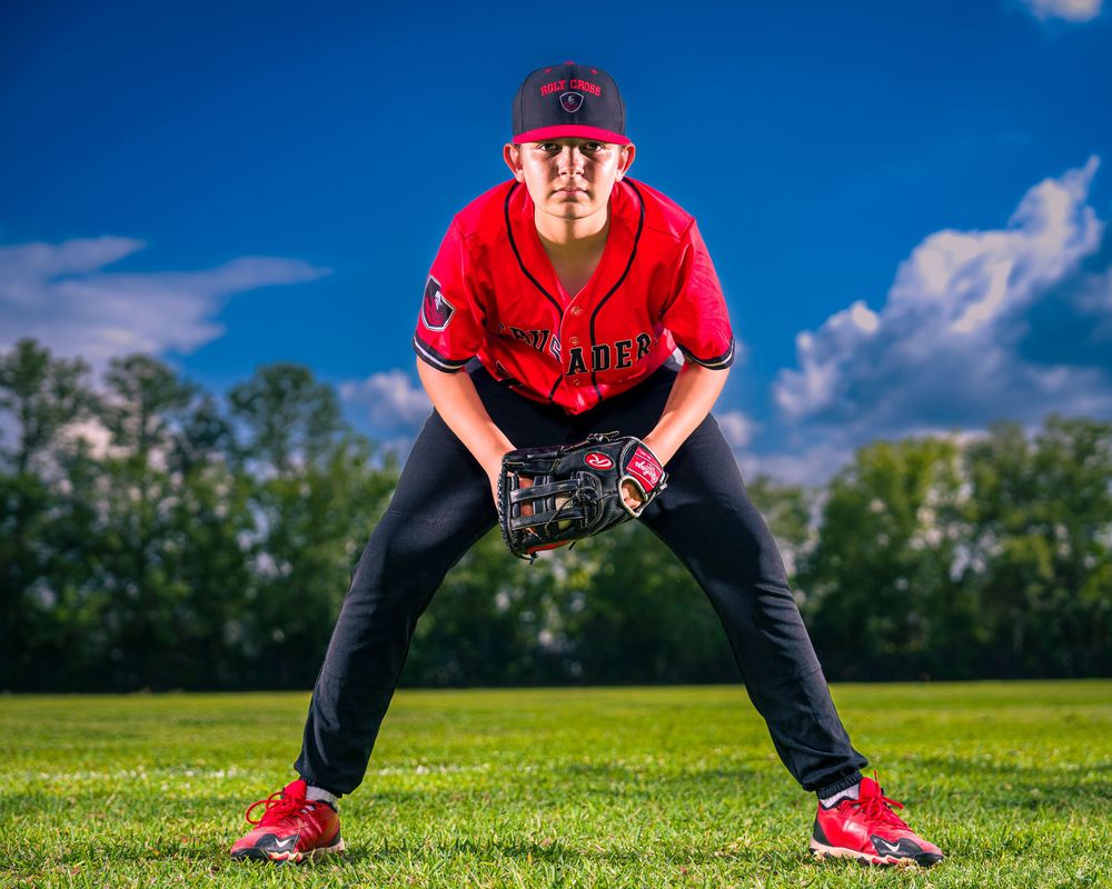 Baseball player in a red jersey and black pants crouches in fielding position on green grass field.