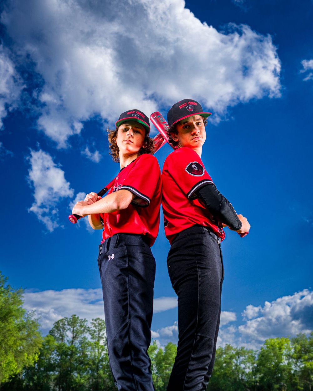 Baseball players in red uniforms standing back to back against a vibrant blue sky with white fluffy clouds.