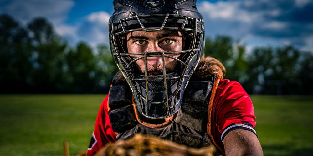 Baseball catcher wearing protective gear and red uniform signals from behind home plate on sunny day at baseball field.
