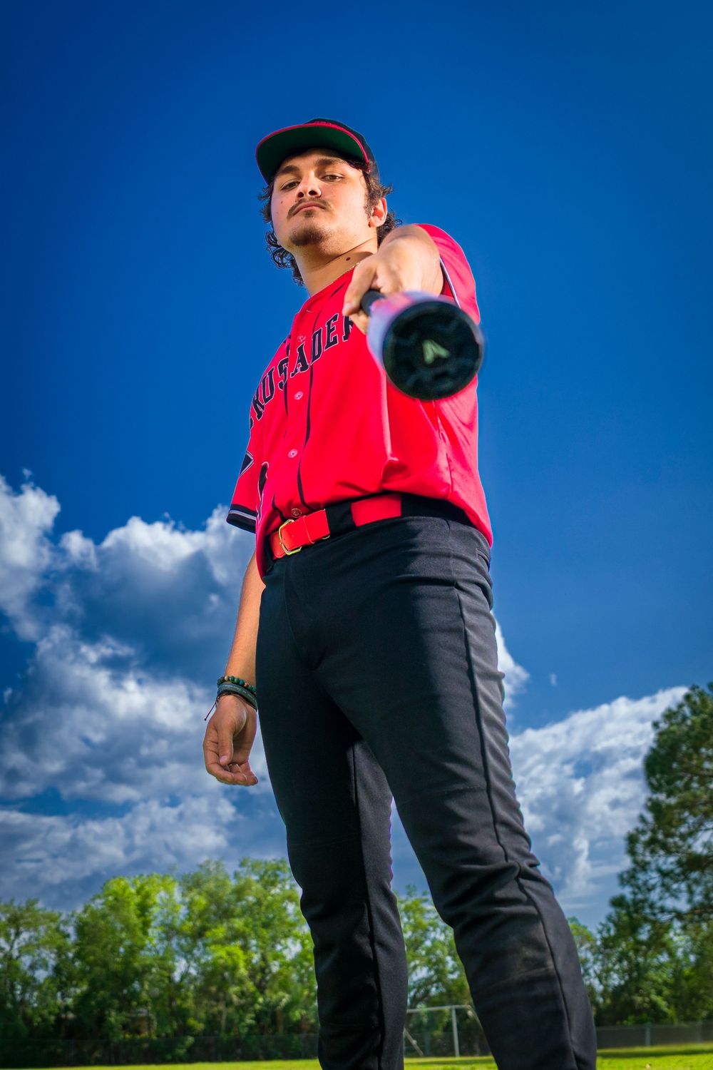 Baseball player wearing red uniform and black cap stands posed against blue sky with white clouds.
