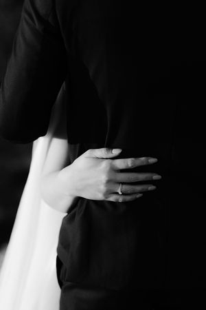 Black and white close-up photo of hands embracing, with a delicate engagement ring visible against formal attire.