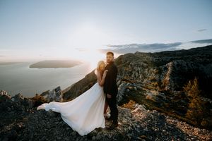 A romantic couple stands on a scenic mountain cliff overlooking a sunset view of the coastline and water below.