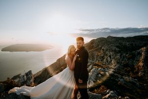 A couple embraces on a rugged mountain overlook with dramatic coastal views and golden sunlight behind them.