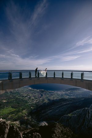 Two silhouetted figures stand on a curved observation deck overlooking a dramatic mountain landscape and blue sea horizon.