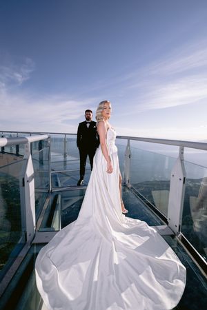 A wedding couple stands on a glass-enclosed balcony overlooking the ocean with a dramatic flowing white bridal gown.
