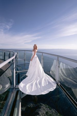 A bride in a flowing white wedding dress stands on a glass-floor observation deck overlooking the ocean and sky.