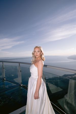 A figure in a flowing white dress poses on a scenic overlook against a dramatic cloudy sky.
