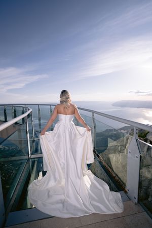 A bride in a flowing white wedding dress admires the scenic mountain view from a glass observation deck.